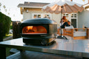 Off Black Gozney Arc Lite with a lit flame on a steel garden table, with friends relaxing behind it.