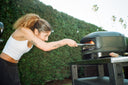 A woman uses a pizza peel in a lit Off Black Gozney Arc Lite on a garden table.