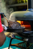 A cook lifts a steaming tomahawk steak beside an Off Black Gozney Dome XL Gen 2.