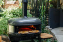 Off Black Gozney Dome XL Gen 2 on a stand with two pizzas on its mantel in a leafy garden.