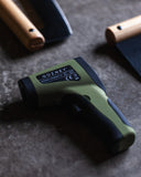 Green and black Gozney infrared thermometer on a stone surface beside wooden-handled pizza rockers.