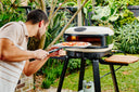 A man loads a pizza into a Bone Gozney Arc XL with a placement peel in a leafy garden.