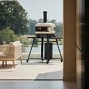 Bone Gozney Dome Gen 2 on a wheeled stand on a sunny terrace overlooking water and trees.