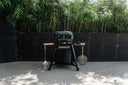 Off Black Gozney Dome Gen 2 oven on a wheeled stand with hanging pizza peels on a bamboo-lined deck.