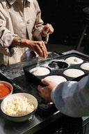 Hands sprinkle flour over dough balls in a black Gozney Dough Tray beside bowls of tomato sauce and cheese.