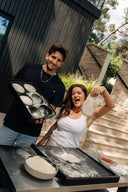 Two people preparing pizza dough with black Gozney Dough Trays at an outdoor table.