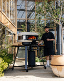 A man tends a wood-fired Bone Gozney Dome Gen 2 on a wheeled stand in a planted courtyard.