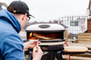 A person uses a turning peel to lift pizza inside a lit Bone Gozney Arc XL on a rooftop terrace.