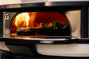 Skillets of broccolini and a baked dish cooking beside flames inside a Bone Gozney Dome Gen 2.