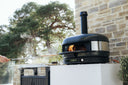Off Black Gozney Dome Gen 2 with a lit flame as a cook holds pizza on a peel beside a stone patio wall.