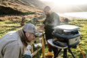 Gozney Tread in Off Black with the Tread Mantel fitted, two people cooking outdoors by the water