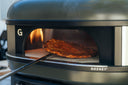 A browned pizza rests on a turning peel inside an Off Black Gozney Dome Gen 2 beside a flame.