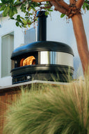 Off Black Gozney Dome Gen 2 with flames inside, framed by a tree and ornamental grasses on an outdoor counter.